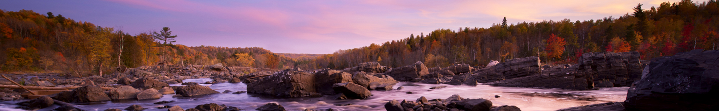 A fast, wide river flowing around huge boulders and through a forest of trees at peak fall color in the mountains of Missouri